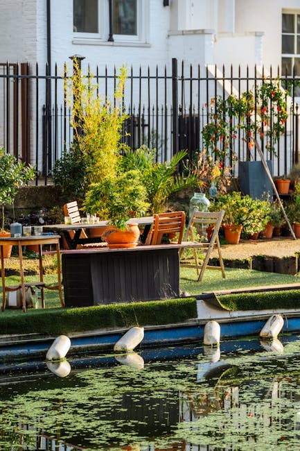 A tranquil garden scene featuring a small canal with floating lily pads and gentle water reflections, bordered by a narrow strip of manicured green grass and a paved pathway. Behind the canal, there is a variety of lush plants and shrubs, including a tall yellow flowering shrub, a potted plant, and leafy green foliage, all situated along a boundary fence made of black metal pickets. In the garden area, outdoor furniture such as a wooden table, chairs, and a small side table are arranged, with some pots and gardening tools visible, indicating ongoing garden maintenance. The setting is illuminated by natural daylight, highlighting the textures of wet soil, trimmed grass, and vibrant plant colors. This scene is captured as part of a garden maintained by Gardeners Catford, aligning with their services in landscaping work and lawn care, as described in the Catford Bridge terrace garden guide SE6.