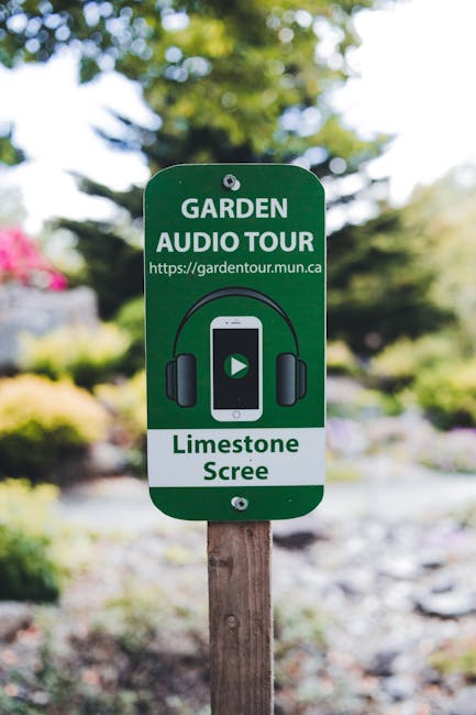 Close-up of a green garden sign mounted on a wooden post, advertising a garden audio tour at Catford Broadway front garden. The sign features a graphic of a smartphone with a play button, surrounded by black headphones, and includes the text ‘GARDEN AUDIO TOUR’ and ‘https://gardentour.mun.ca’. Below, it specifies ‘Limestone Screen’. In the background, blurred trees with green leaves, shrubs, and flowering plants are visible, indicating a landscaped outdoor setting with a mix of lawn areas, flower beds, and pathway surfaces. The scene is lit by natural daylight, highlighting the lush greenery and well-maintained garden environment. Gardeners Catford specializes in garden maintenance and landscaping work, ensuring this outdoor space remains vibrant and inviting.